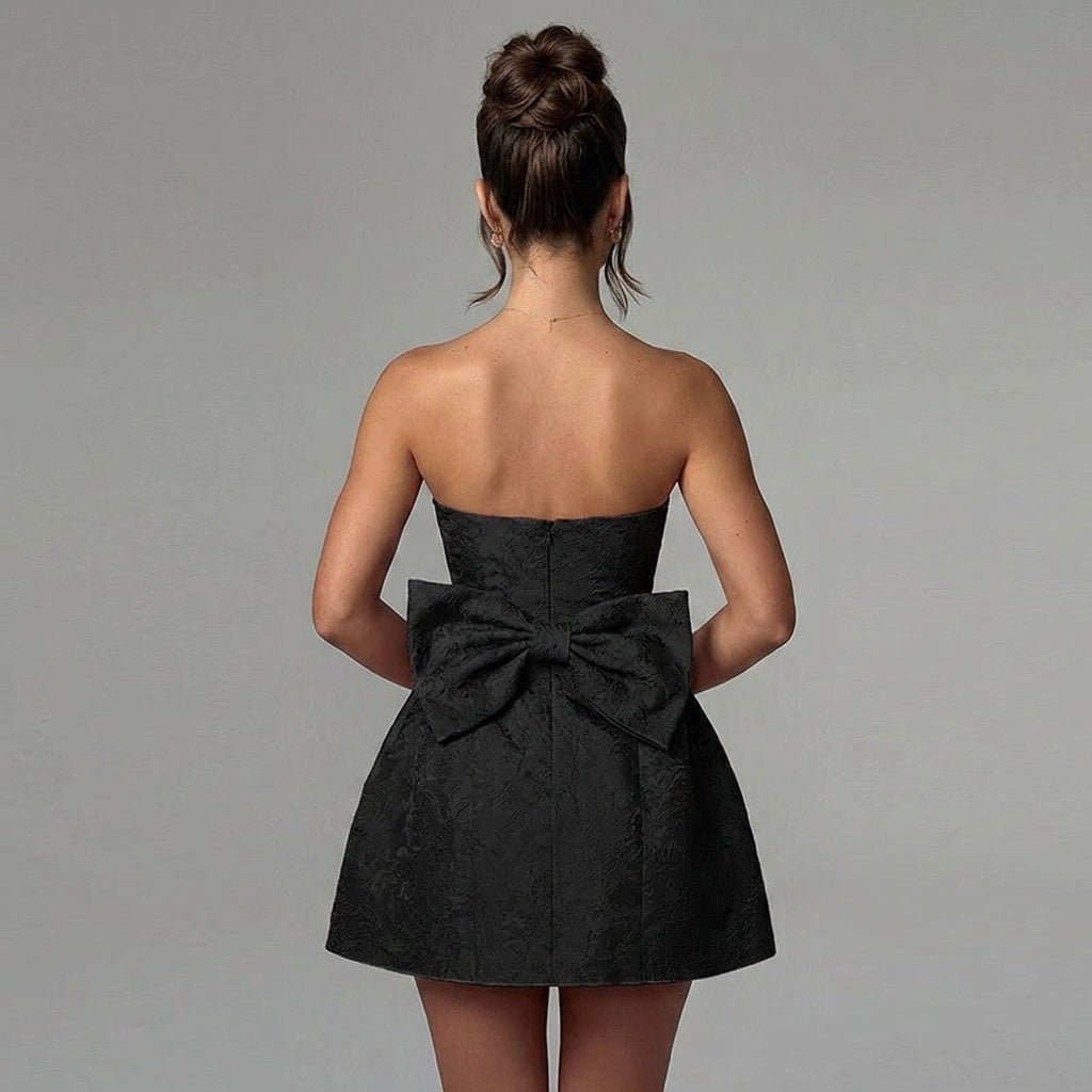 Back view of female model wearing a black mini dress featuring a large statement bow and floral textured fabric by Kouvr.
