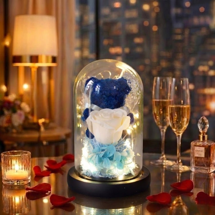 Blue moss teddy bear with a white rose in a glass dome featuring a sleek black base and warm white lights.