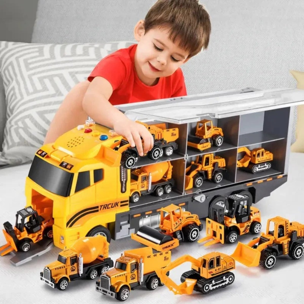 A young boy in a red shirt smiling as he places a small construction vehicle into the storage compartment of the large yellow Kouvr carrier truck toy on his bed.