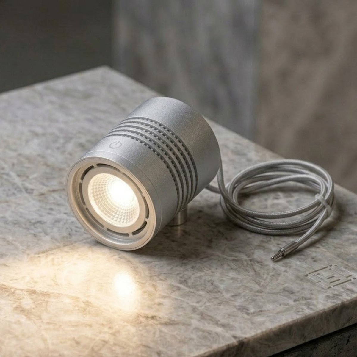 A close-up view of a small, cylindrical aluminum LED light puck. The light is made of silver metal with a power symbol on top and cooling ridges. It is illuminated, casting light on a light-colored marble surface. A grey fabric-wrapped electrical cord is coiled next to it, with a metal tip plug visible.