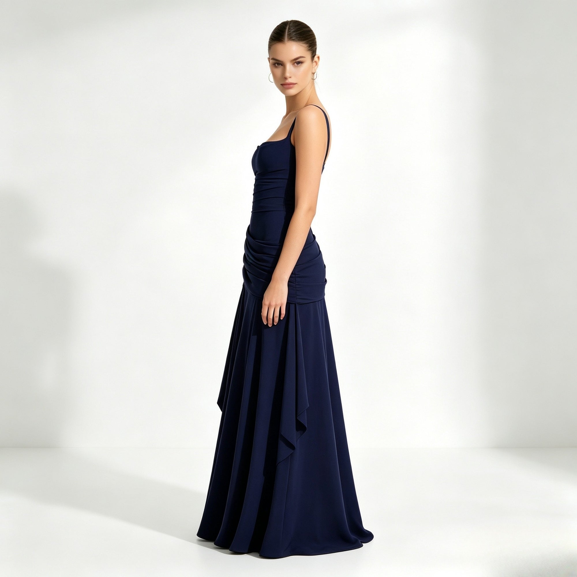 Side profile of a woman standing in a dark blue floor-length flowy formal maxi dress featuring structured asymmetrical draped hip details.