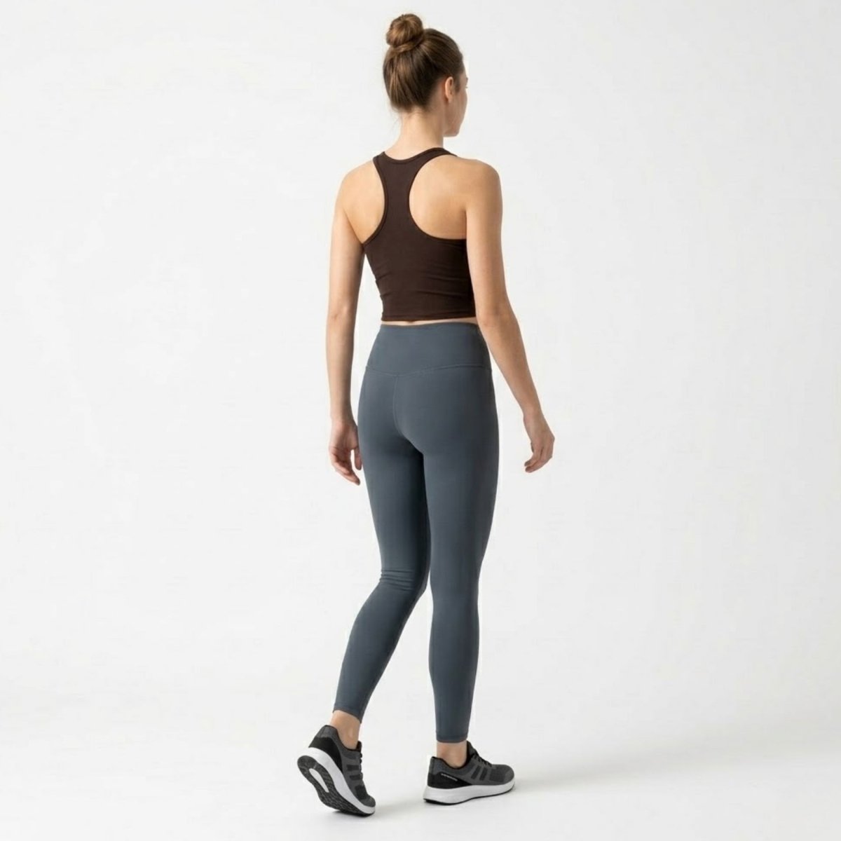 Full body back view of a woman wearing a dark brown racerback sports tank and grey high-waisted leggings.
