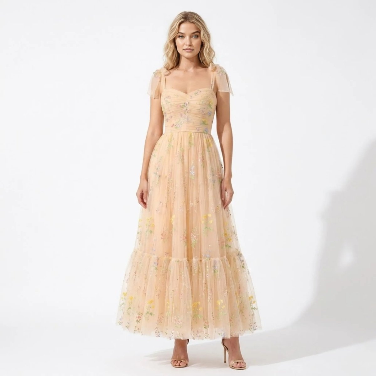 Woman wearing Kouvr floral embroidered tulle maxi dress in champagne featuring tie shoulder straps and sweetheart neckline.