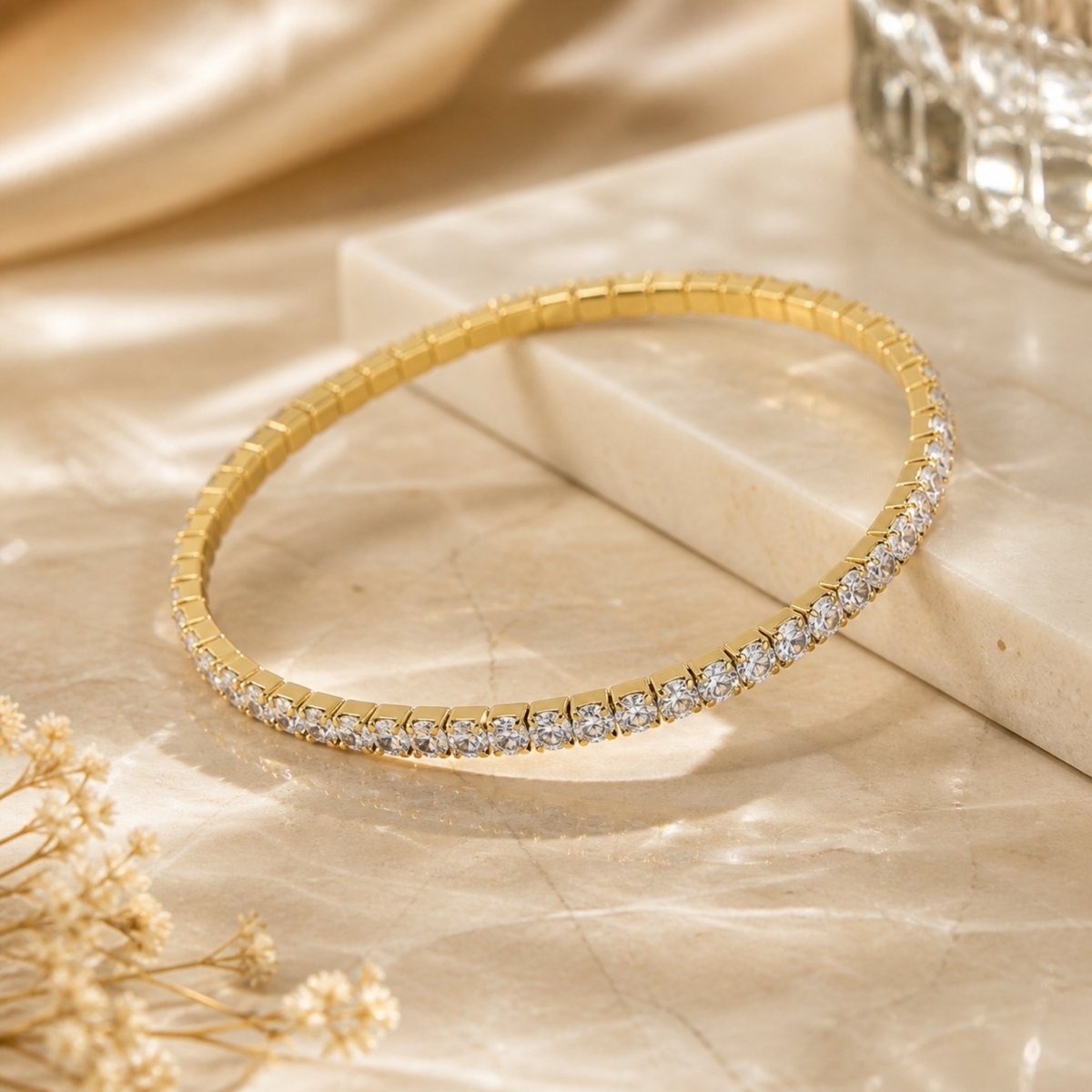 Aesthetic golden hour product photography of the gold plated slip-on 3mm tennis bracelet resting on marble tile with floral props and dramatic sunlight shadows.