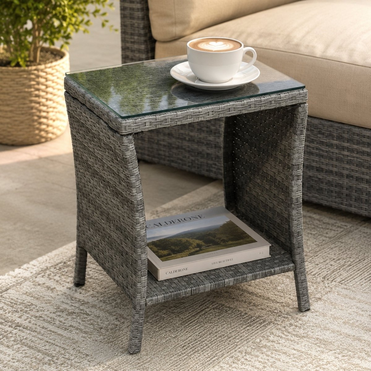 Luxury grey wicker outdoor side table with a clear glass top and open lower shelf, styling a coffee cup and design book on a patio.