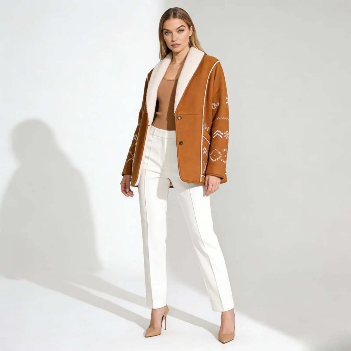 Model wearing the Kouvr Alice embroidered shearling jacket in caramel brown, paired with white trousers.