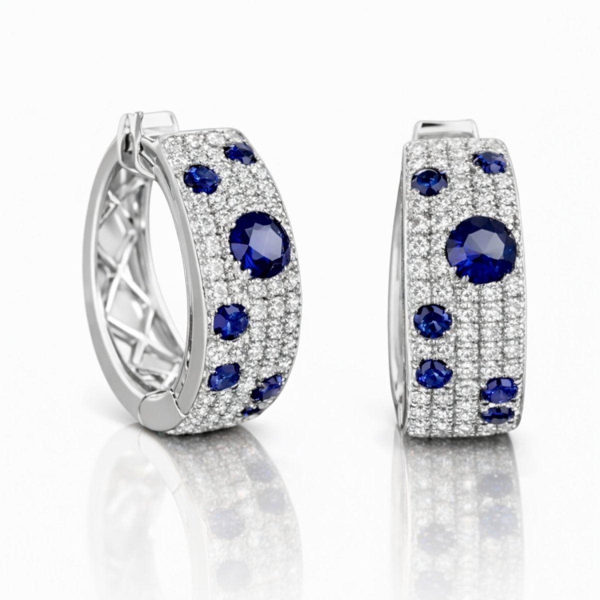 A pair of Kouvr Amaris hoop earrings with pavé diamonds and round blue sapphires in 18k white gold on a reflective white background, showing the front and inner gallery.