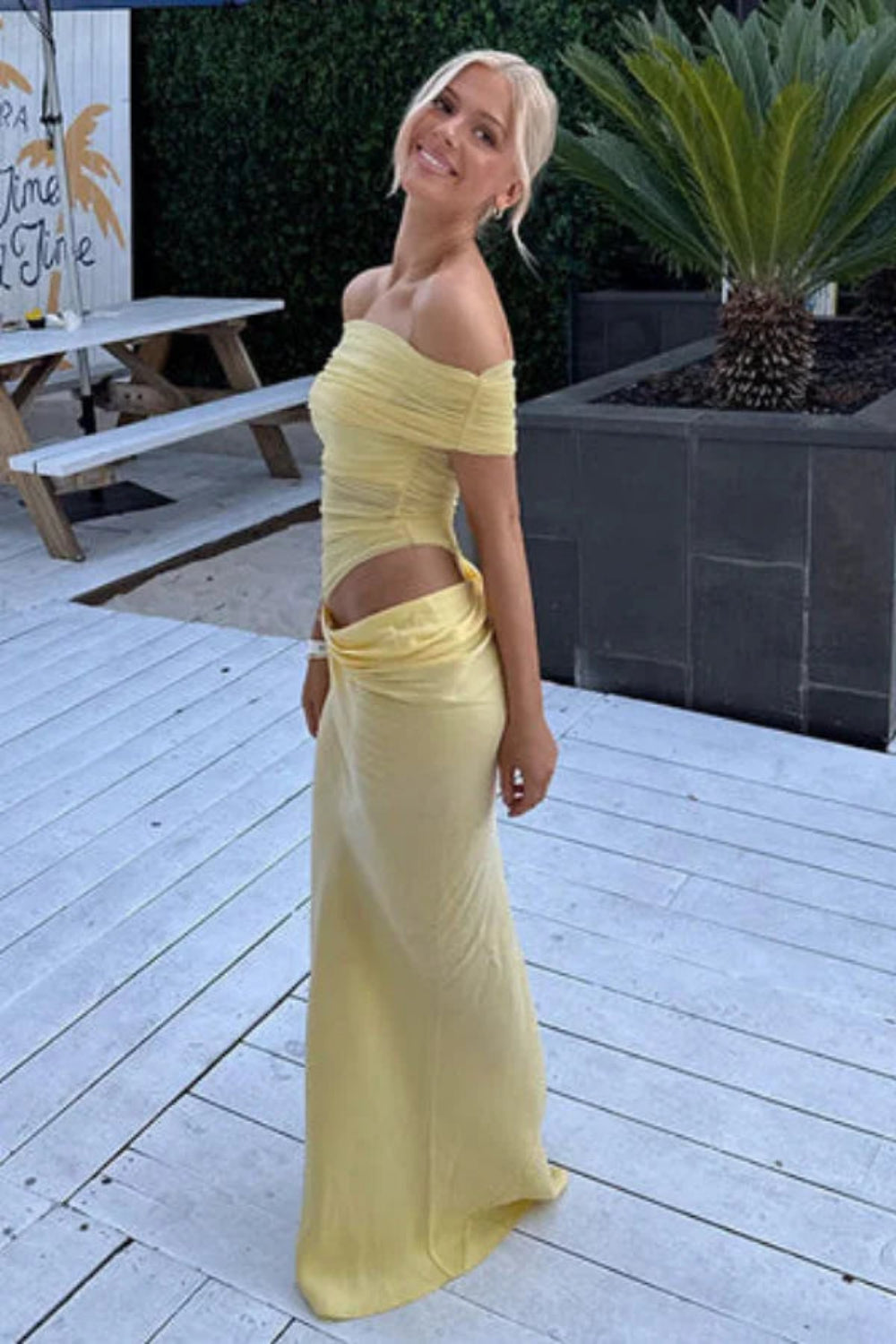 Model wearing the Kouvr Anita Gown in Lemon Chiffon, a yellow off-the-shoulder maxi dress with a side waist cutout