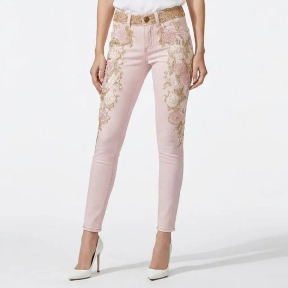 Zoom in to jean. Female model facing forward wearing Kouvr Beth blush pink skinny jeans with intricate gold and white floral embroidery, styled with a white t-shirt and white heels.