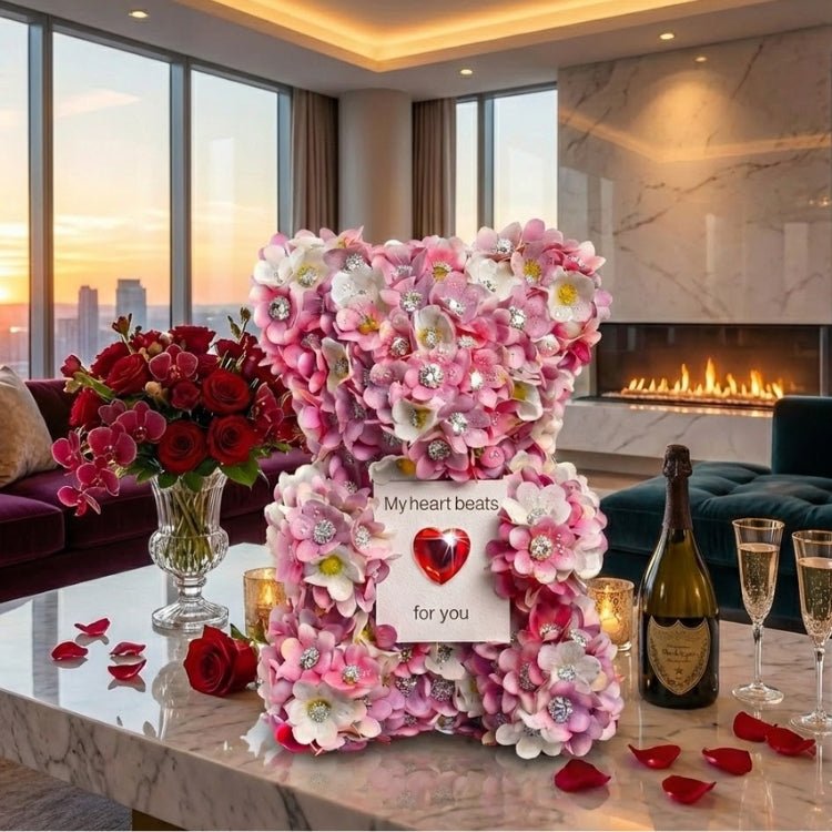 The Kouvr Blossom & Jewel Bear made of pink and white flowers on a table in a luxury penthouse overlooking a sunset skyline with champagne.