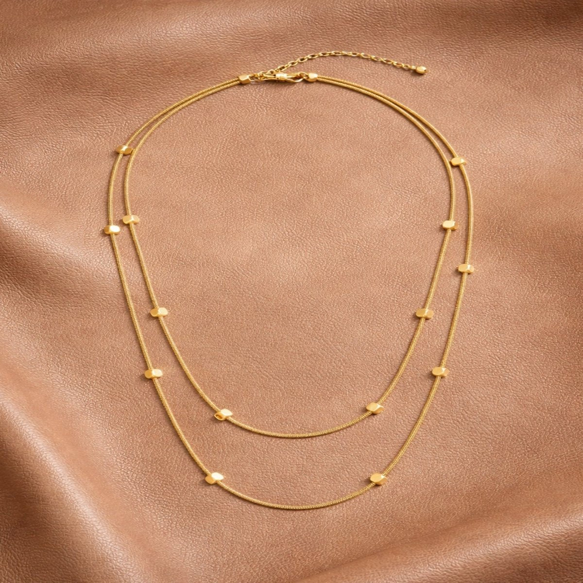 Full view of the Kouvr Celine double layer 18k gold plated necklace laid flat on a brown leather surface.