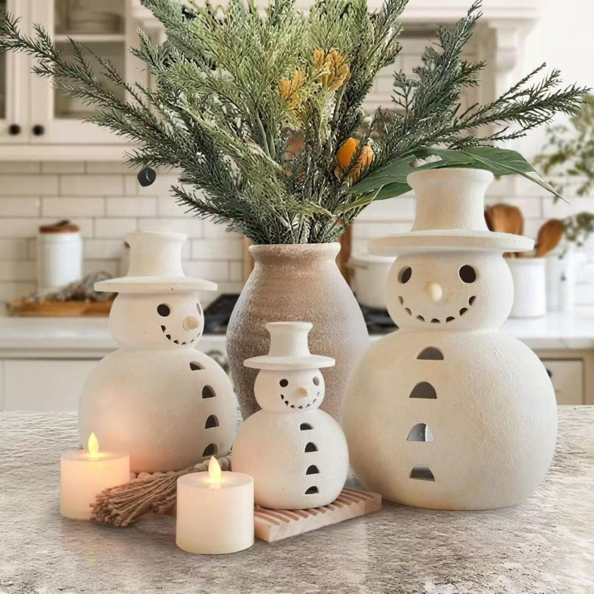 A set of three white ceramic snowman luminaries of varying sizes arranged as a centerpiece on a rustic table, glowing warmly with LED candles beside a vase of evergreen branches.