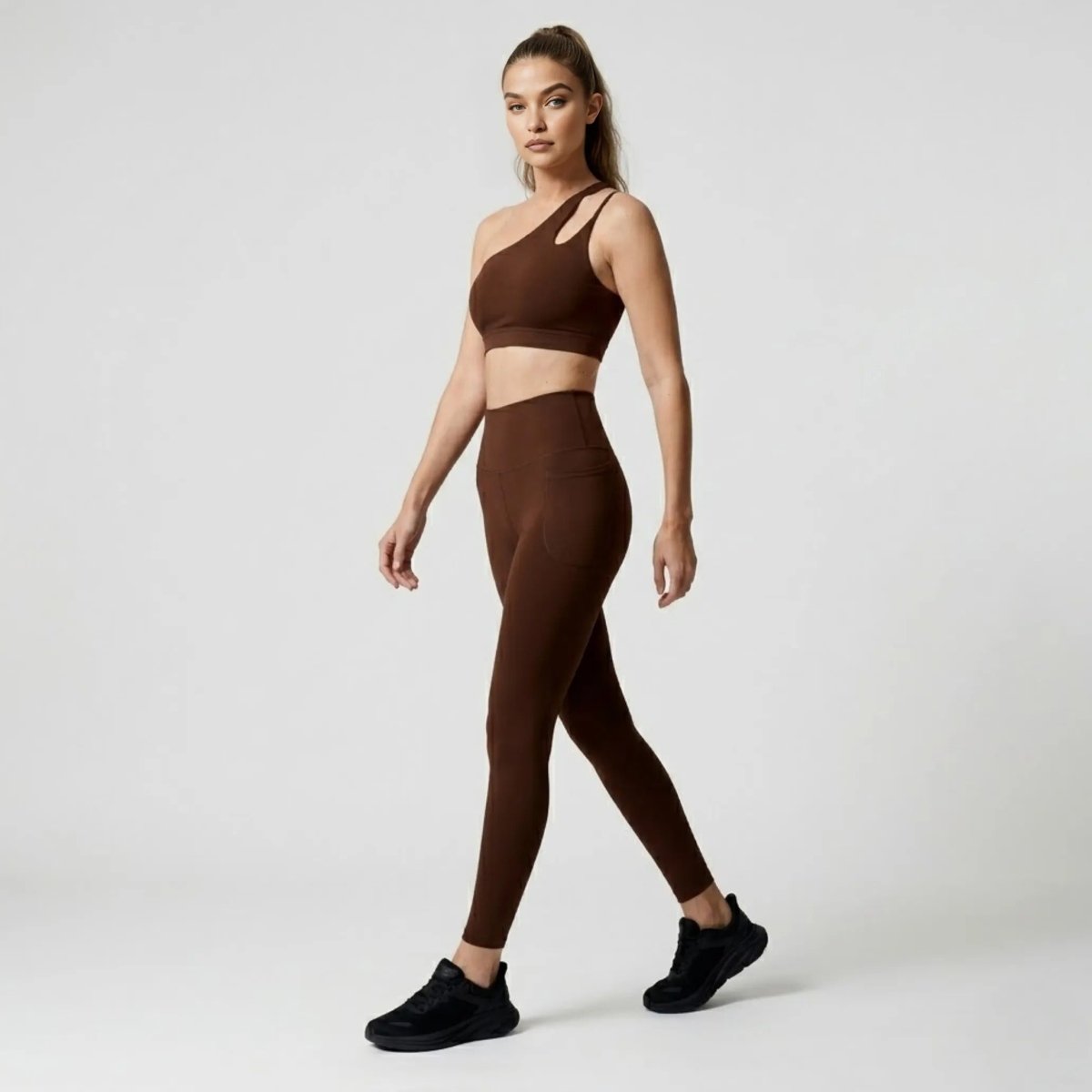 Side profile of model walking in luxury affordable dark brown high waisted workout leggings and asymmetrical sports bra by Kouvr.