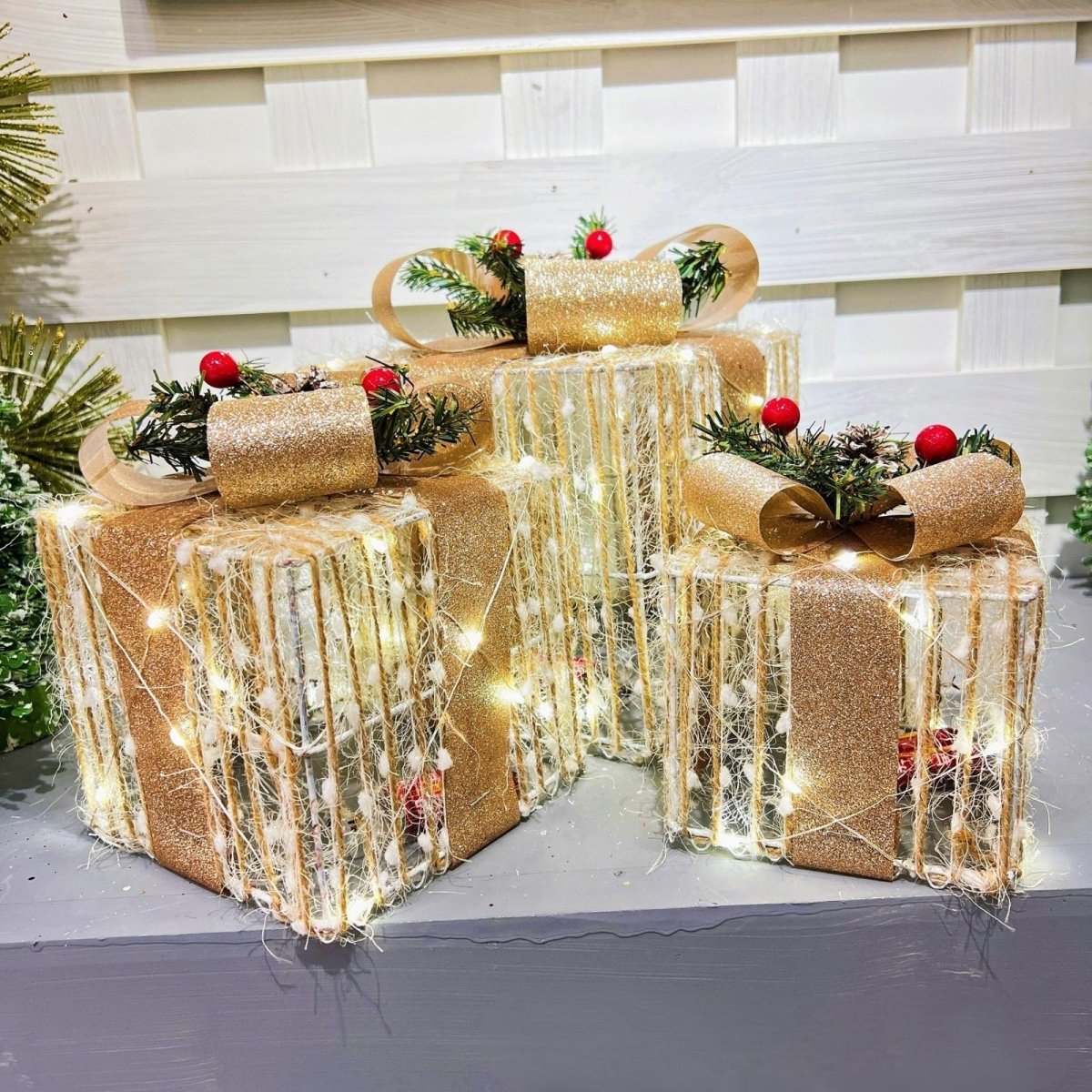 A close-up view of the Kouvr Éclat set of three lighted gift boxes, showing the glitter, twine, and warm LED light detail against a white shiplap wall.