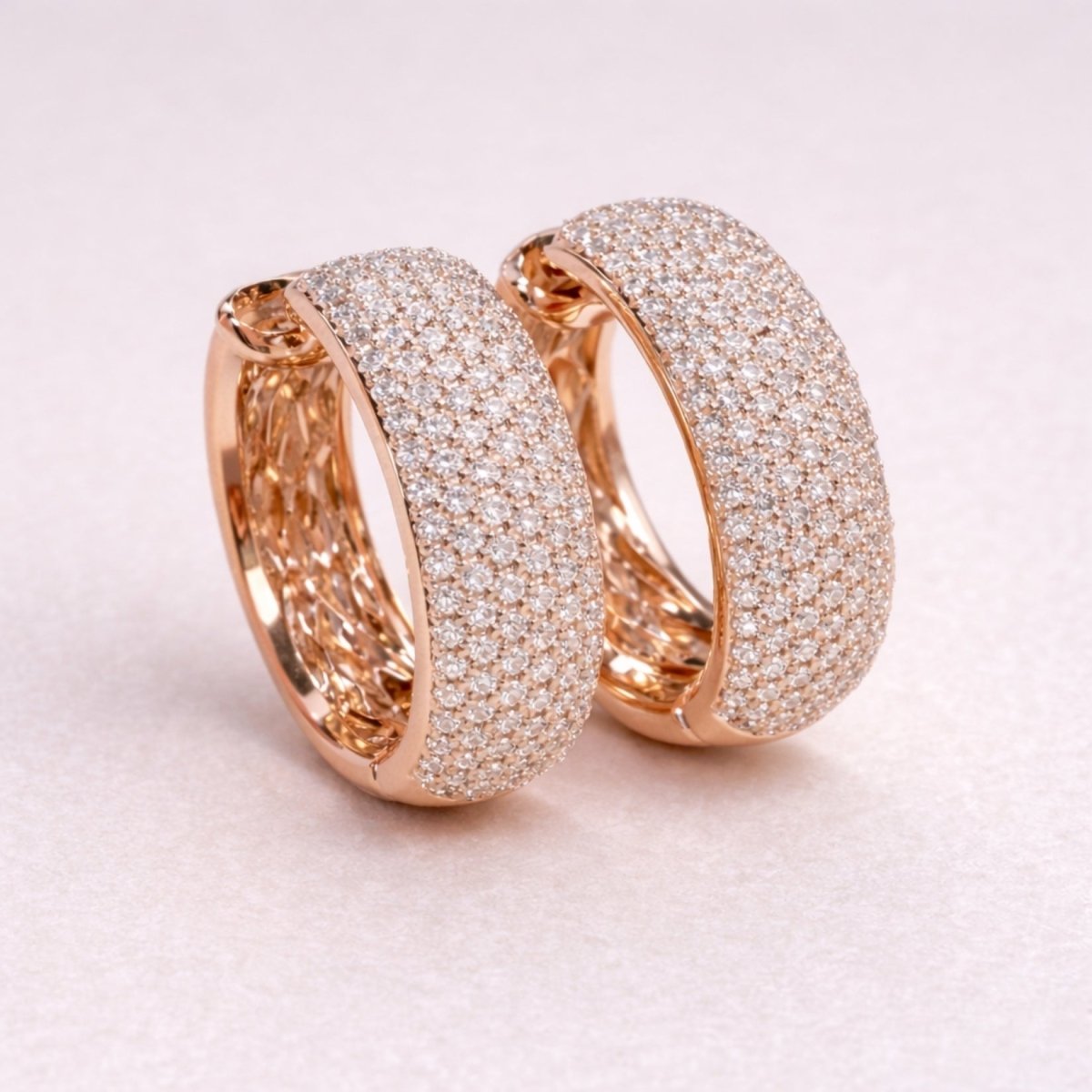 A pair of Kouvr Emmiline wide hoop earrings in 18k rose gold, fully encrusted with pavé diamonds, sitting on a light cream surface.