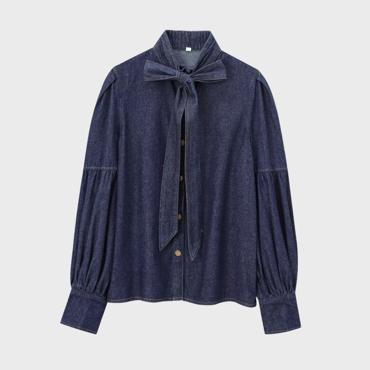 Flatlay product shot of the Kouvr Fashion dark wash denim blouse, with the Alberta-bow tied and buttons fastened, against a light grey background.