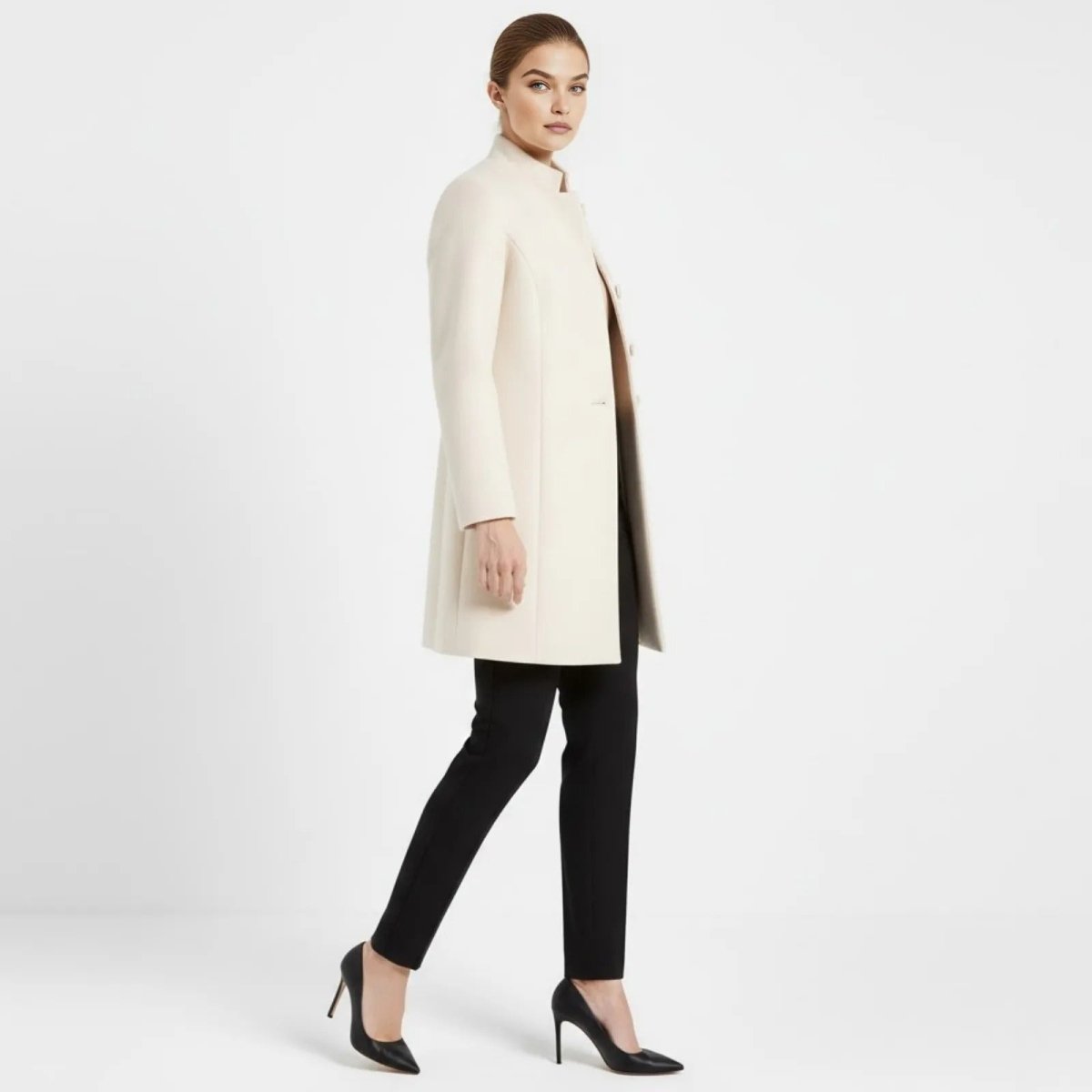 Side view of a model walking in the cream wool A-line coat by Kouvr Fashion, showcasing its silhouette and mid-thigh length.