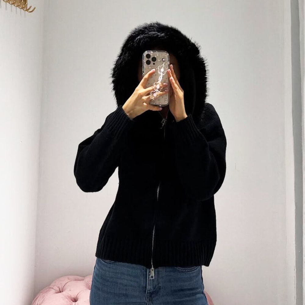 Person wearing a black kouvr hoodie with fur trim, holding a phone in front of their face.