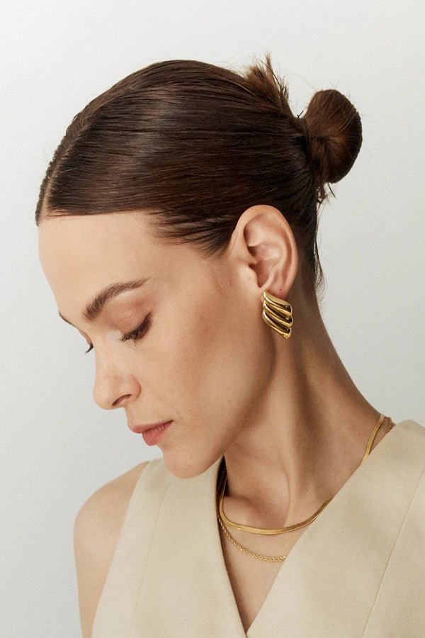 A model with her hair in a bun wears the Kouvr Fashion gold sculptural wavy statement earrings, showcasing their profile against her skin.