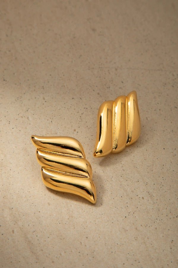 A product shot of a pair of Kouvr Fashion gold wavy statement earrings laid on a textured neutral surface.