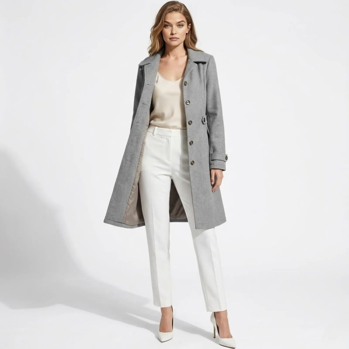 Woman wearing the Kouvr Fashion Priscilla grey wool coat open with a cream top, white trousers, and white heels, standing against a white background.