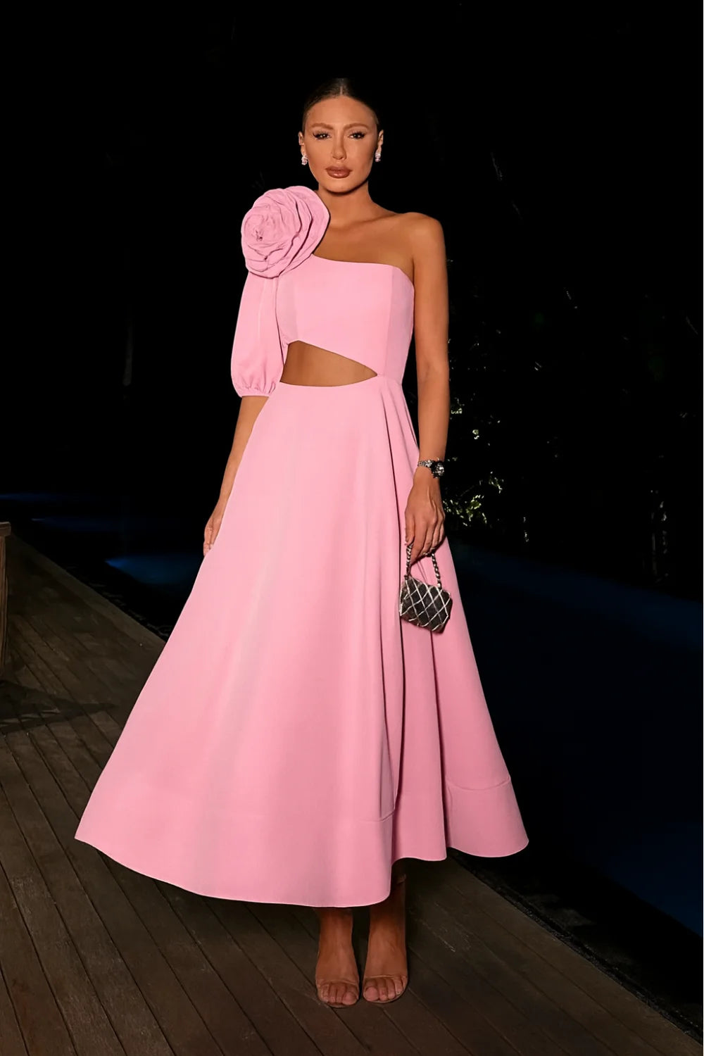Model wearing Kouvr's pink one-shoulder midi dress with a large rose on the shoulder and a side waist cutout, standing by a pool at night