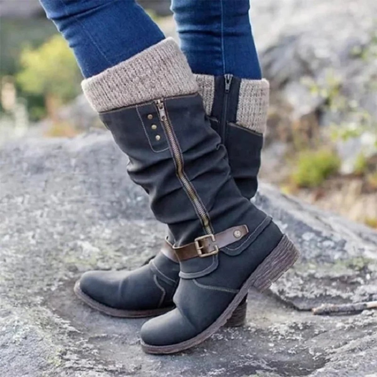 A woman wearing Kouvr Fashion's black laura knee-high boots with knit cuffs and blue jeans, standing on a large rock outdoors.