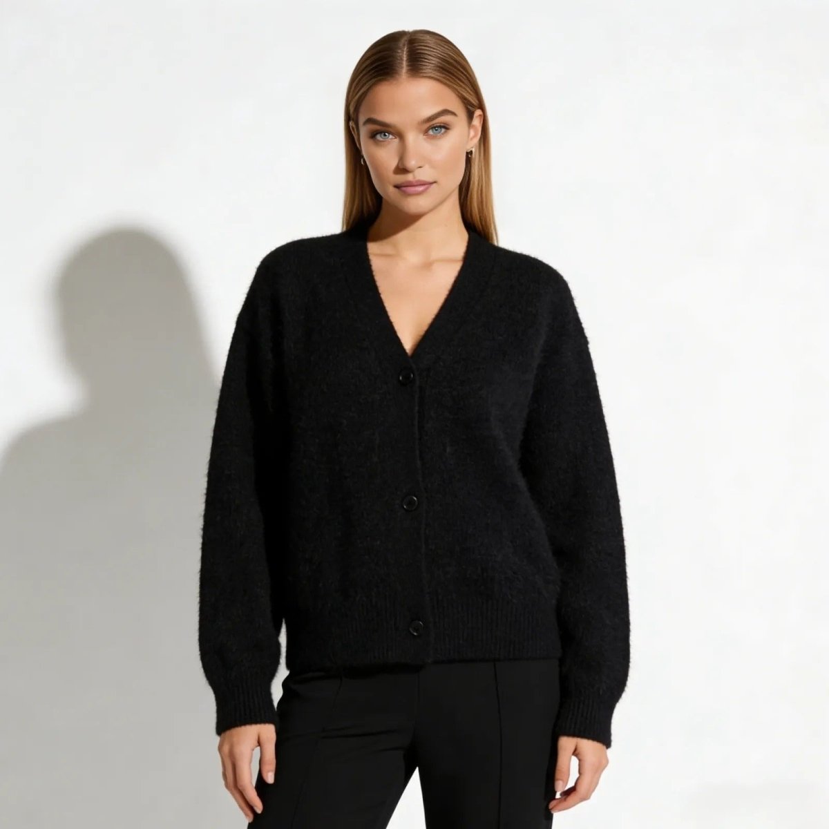 Model with blonde hair wearing the Kouvr Gina black fuzzy V-neck cardigan buttoned up, paired with black trousers, against a white studio background.