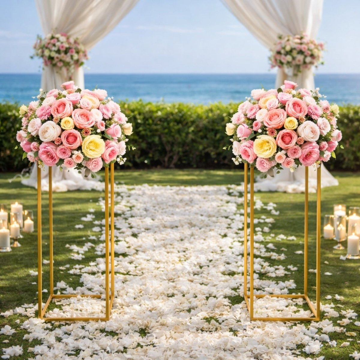 A pair of gold geometric centerpiece stands with pink and yellow rose arrangements framing a wedding aisle on a green lawn overlooking the ocean.
