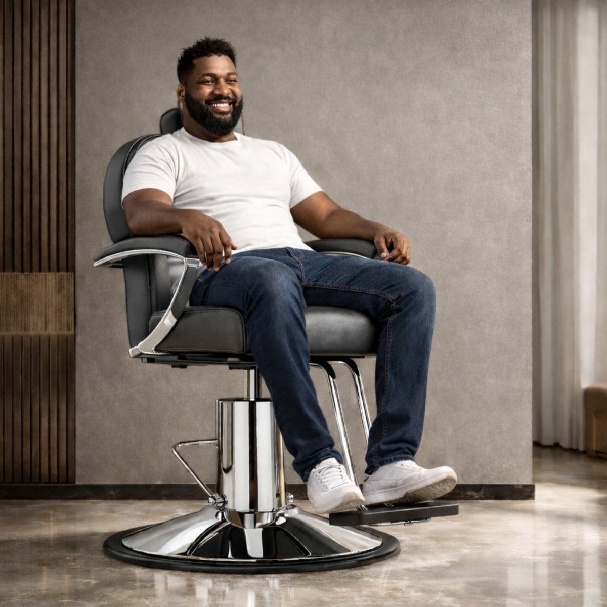 A smiling man sitting comfortably in a black leather barber chair supported by the Kouvr heavy-duty chrome hydraulic pump base.