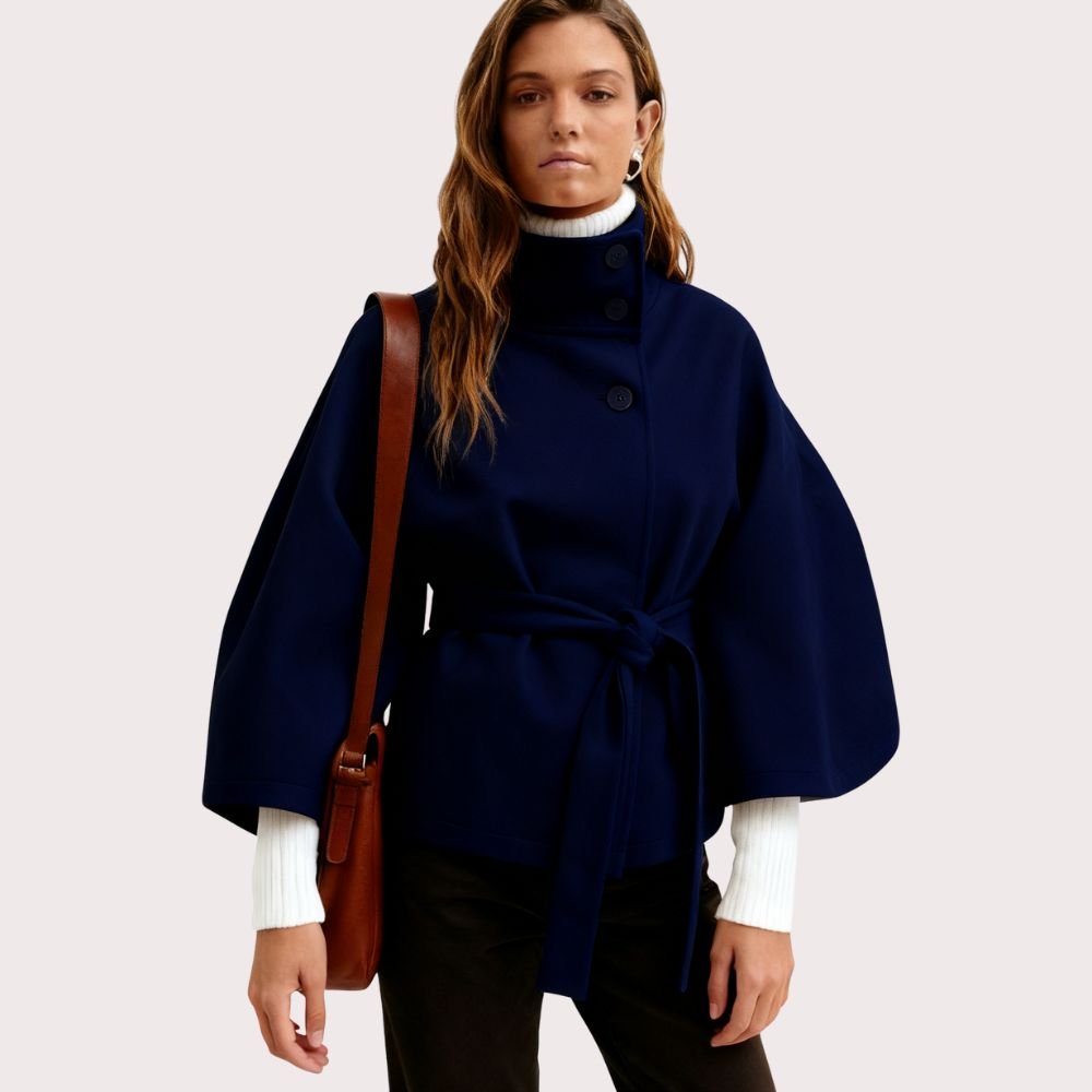 Woman wearing the Kouvr Jaye Cape in navy blue, featuring a high funnel neck, wide sleeves, and a self-tie belt