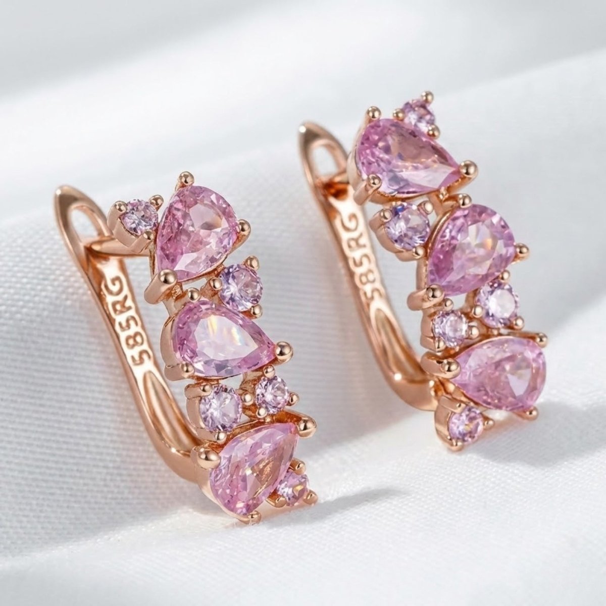 A pair of Kouvr Laliana 14K rose gold huggie earrings featuring a cluster of pear-shaped and round pink sapphire gemstones on a white silk background.
