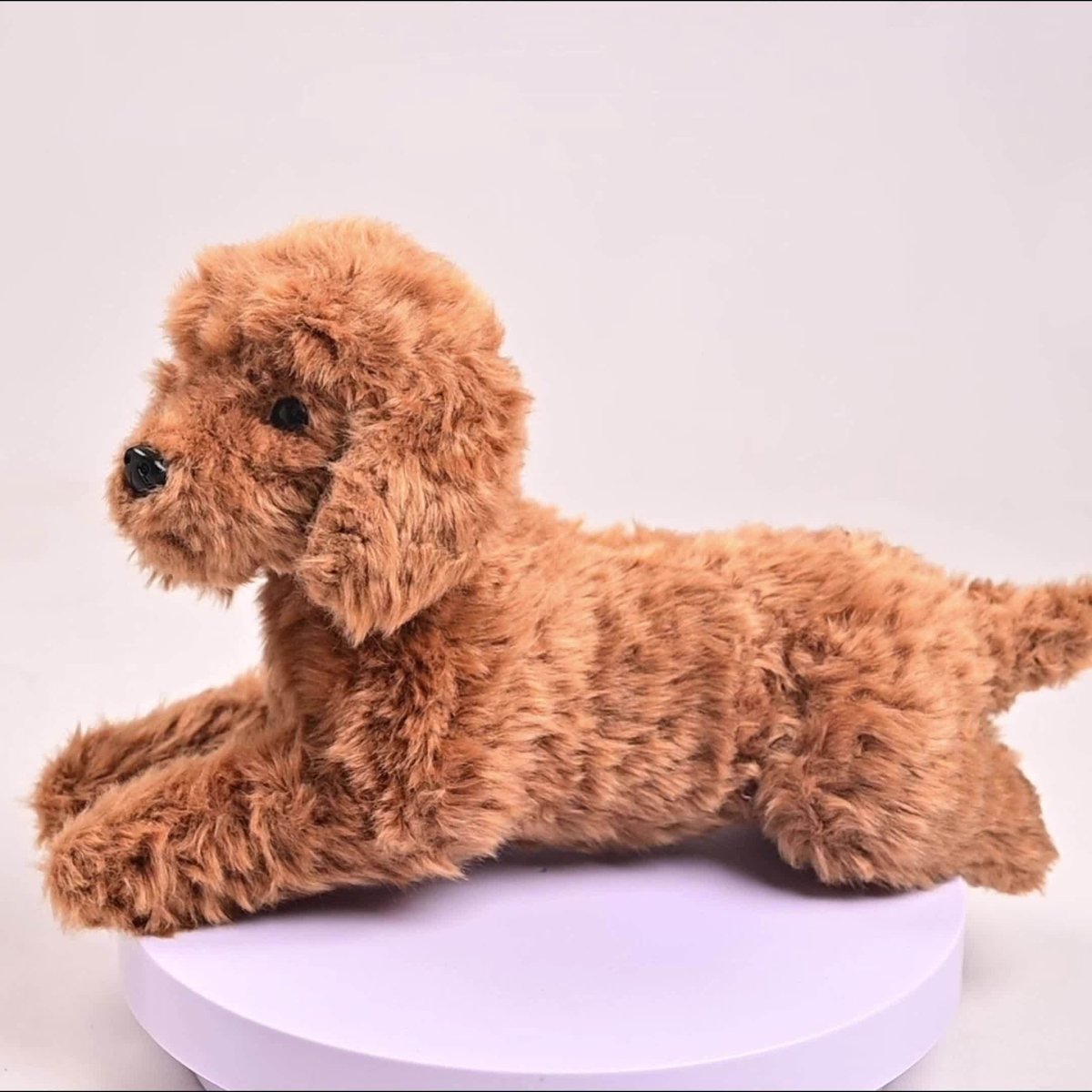 Side view of the Kouvr realistic brown poodle plush toy lying down, showcased on a light purple display stand against a white background.