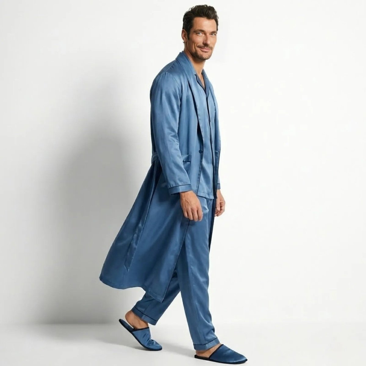 Man smiling while walking in a Kouvr azure blue satin robe and pajama set, showing fabric movement.