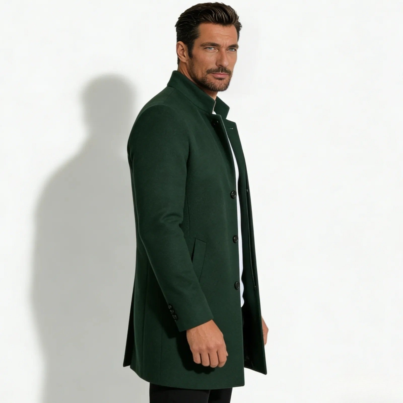 Side profile of a man in a slim fit dark green wool topcoat with a stand collar, showing the coat's silhouette and length.