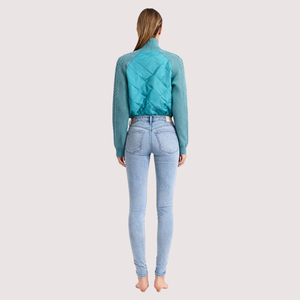 Model wearing the Kouvr Mika Hybrid Bomber Jacket in Aegean Teal, featuring cable-knit sleeves and a diamond-quilted body, paired with light-wash skinny jeans and white sneakers