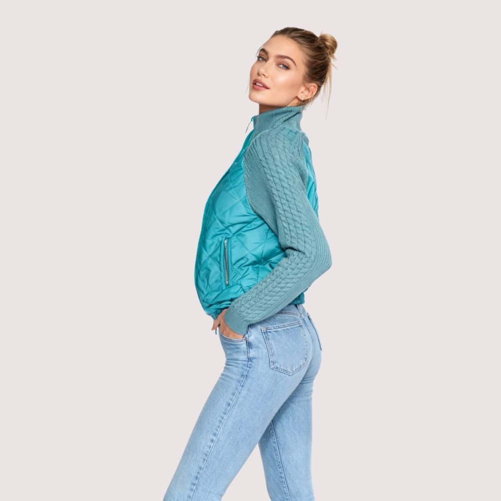 Model wearing the Kouvr Mika Hybrid Bomber Jacket in Aegean Teal, featuring cable-knit sleeves and a diamond-quilted body, paired with light-wash skinny jeans and white sneakers