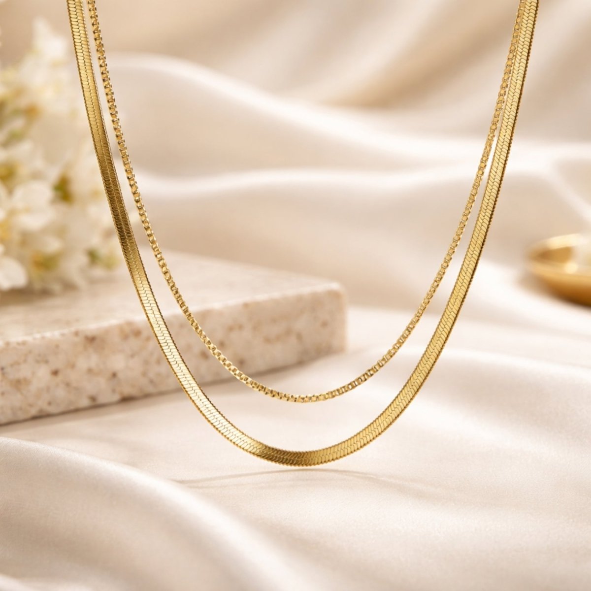 Close-up detail shot of the Kouvr layered gold necklace on a cream satin background, showing the texture of the snake and box chains.
