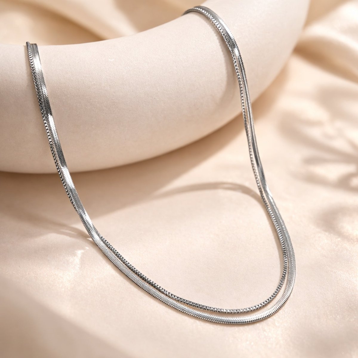 Close-up detail of the Kouvr silver herringbone and box chain layered necklace draped over a textured beige display stand.