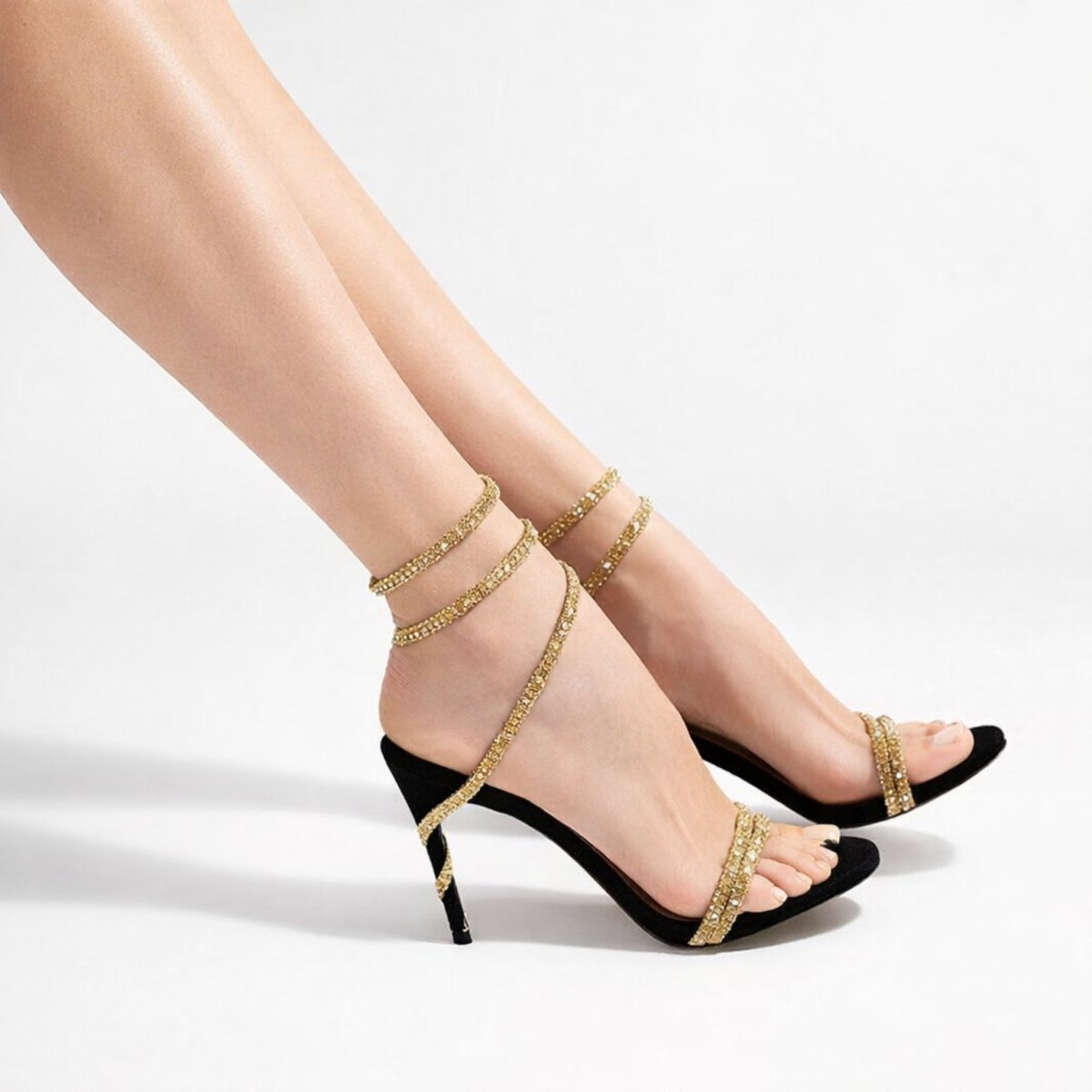 Side profile of a woman's feet wearing black high-heeled sandals with golden crystal straps that spiral up the ankle and heel.