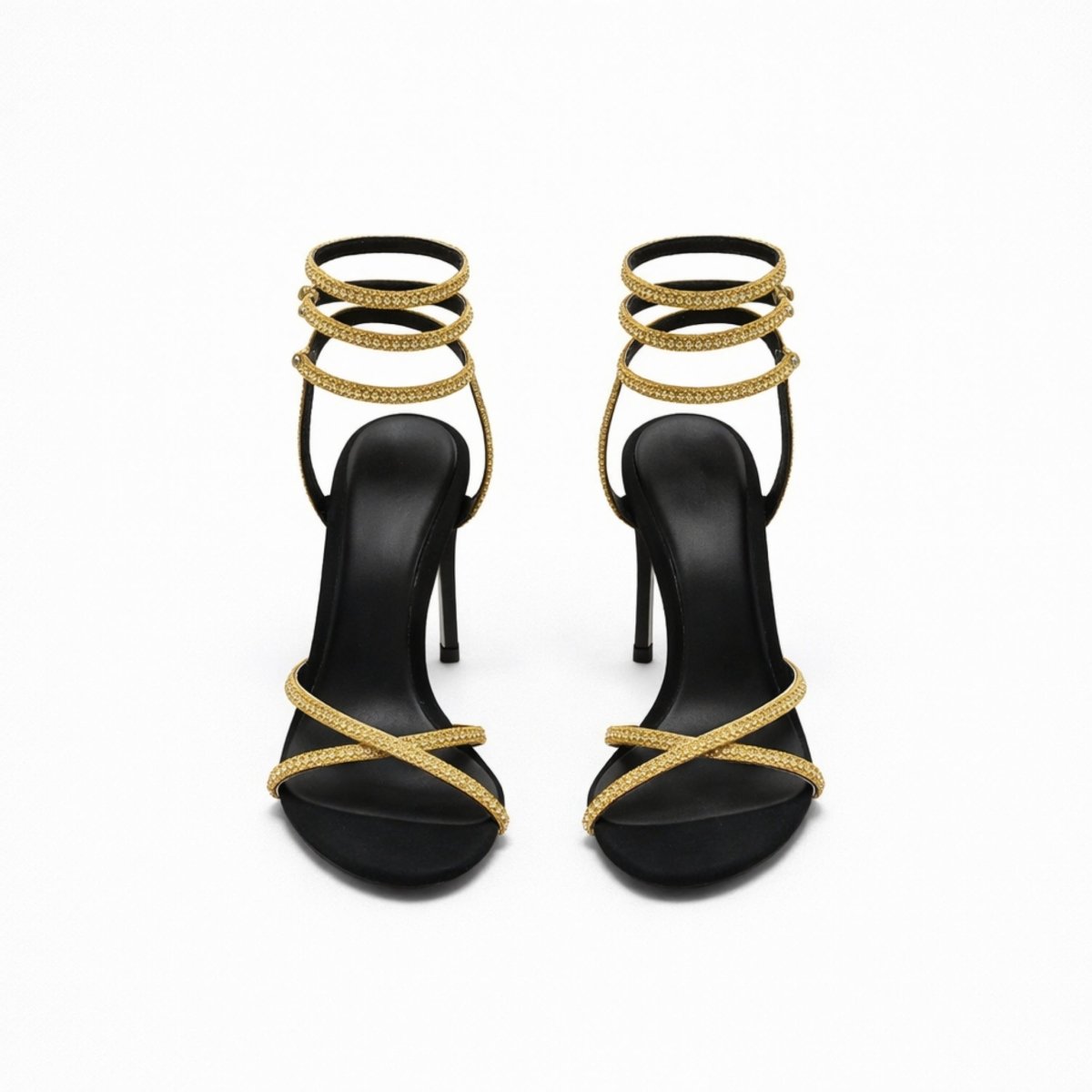 A pair of Kouvr Mireya black stiletto sandals with gold crystal serpent straps wrapping around the ankle, shown from the front on a white background.