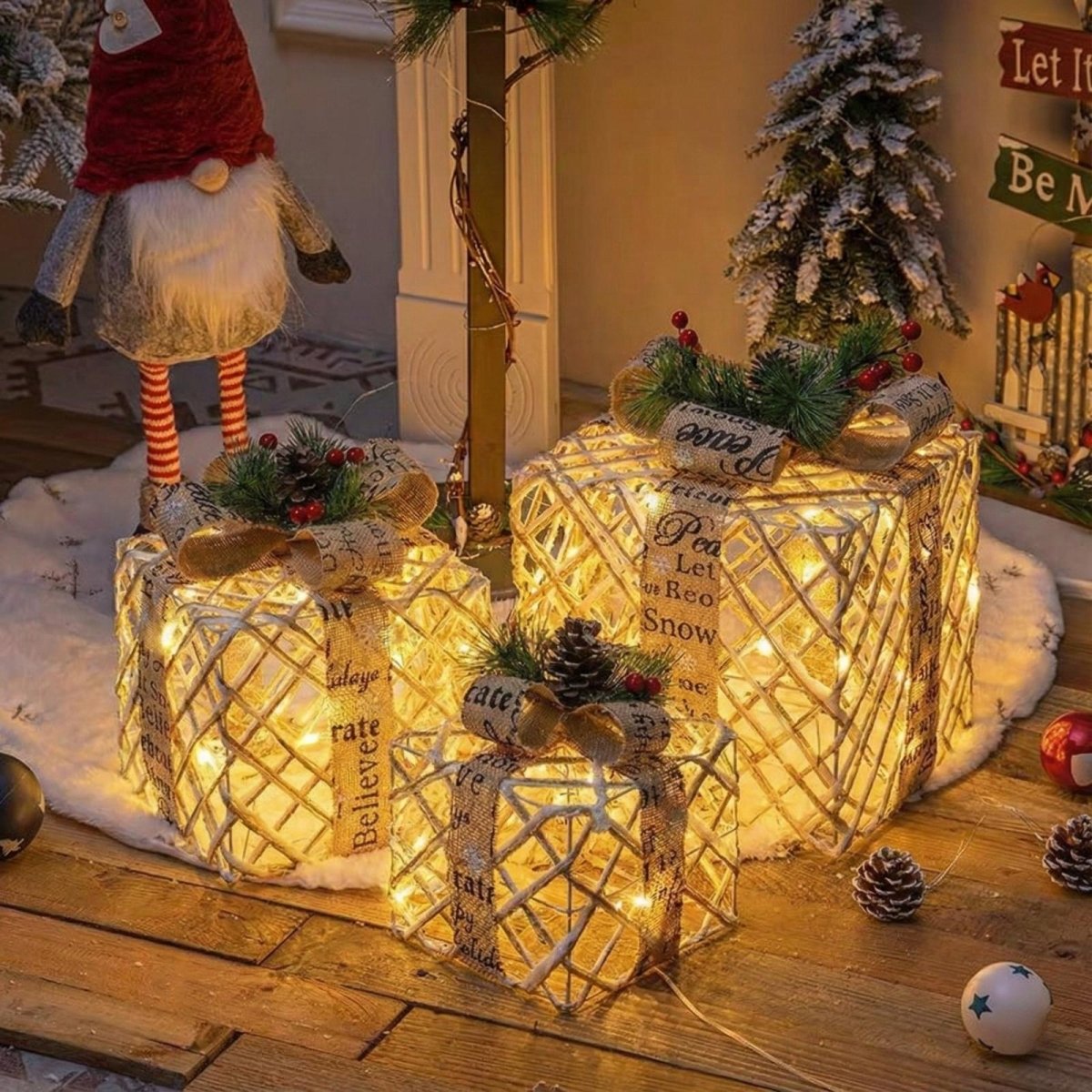 Set of three gold glitter lighted Christmas gift boxes with bows sitting on a white tree skirt under a Christmas tree next to a gnome decoration.