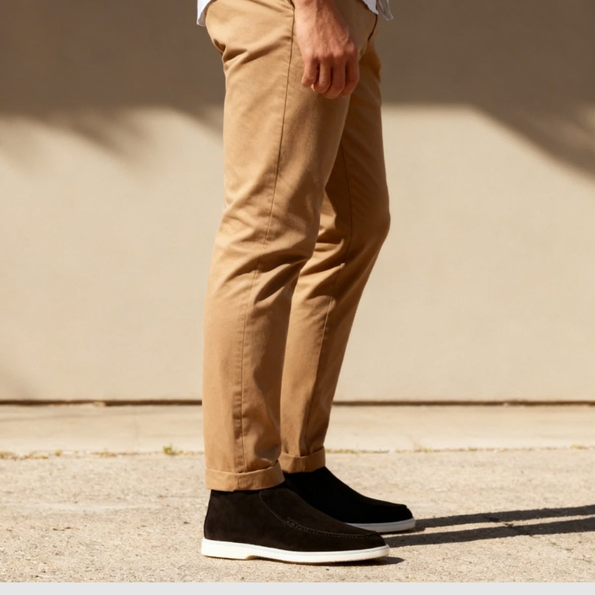 Side view of the Kouvr Ralph black suede chukka sneaker, highlighting the slip-on design and contrast white sole with cuffed tan trousers.