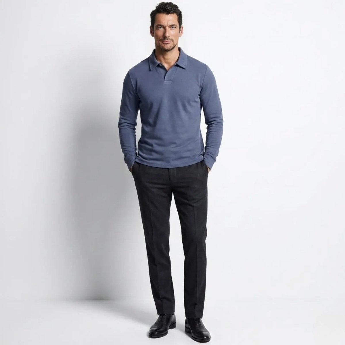 Man wearing the Kouvr Rome Long Sleeve Polo in slate blue, standing front-on with hands in pockets of dark tailored trousers.