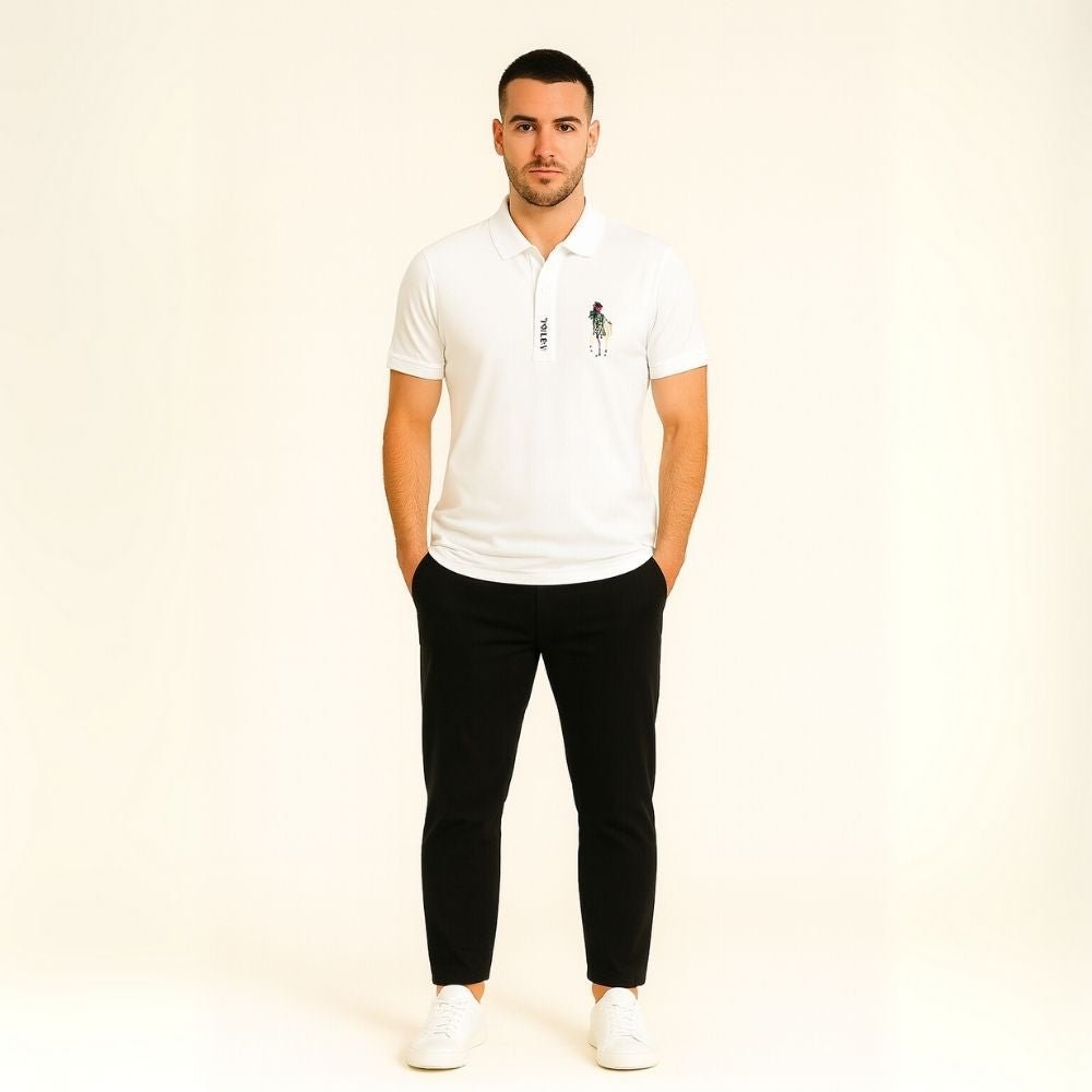 Man wearing a white kouvr polo shirt with a small graphic and black pants on a beige background