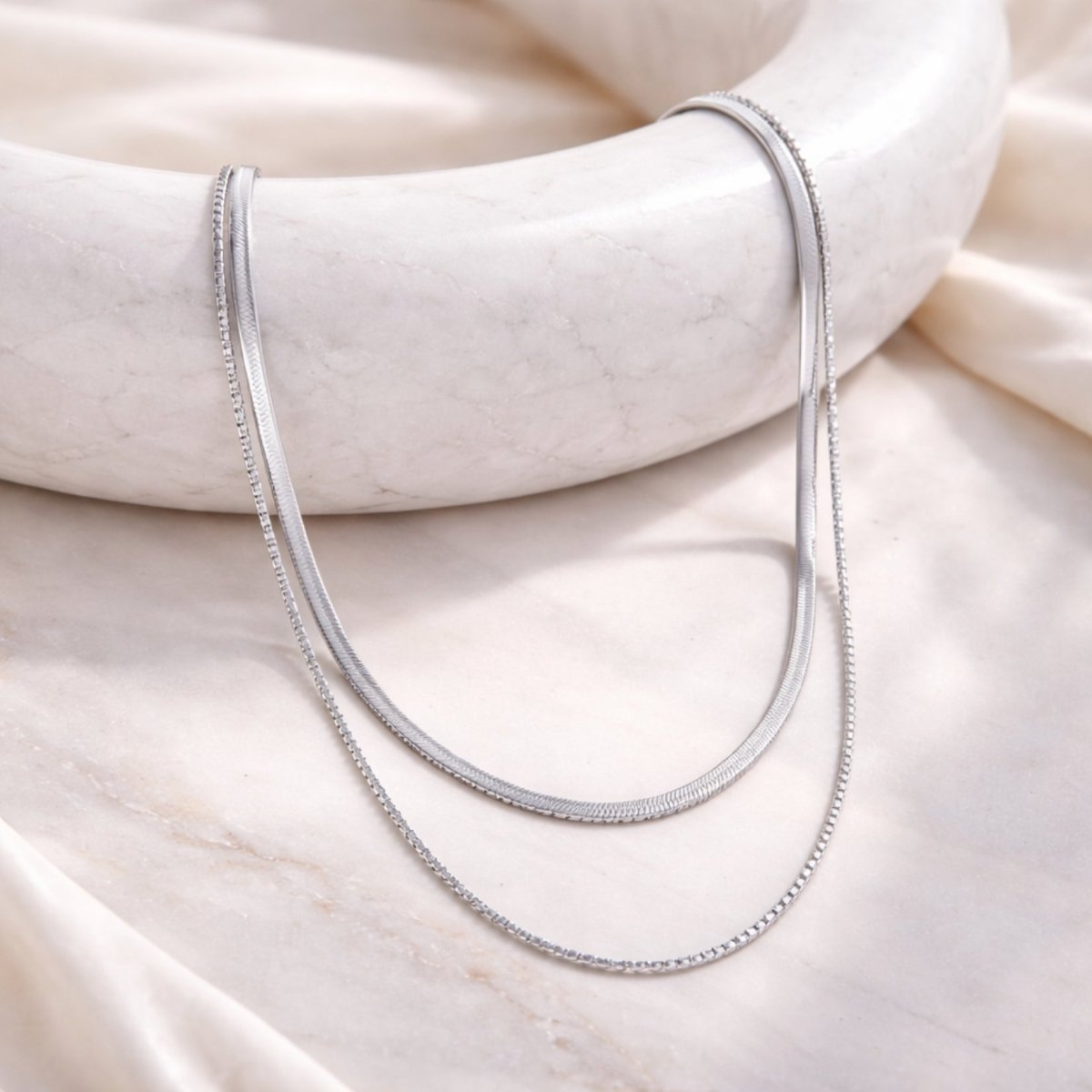 The Kouvr dual-chain silver necklace displayed elegantly on a white marble surface with a silk background.