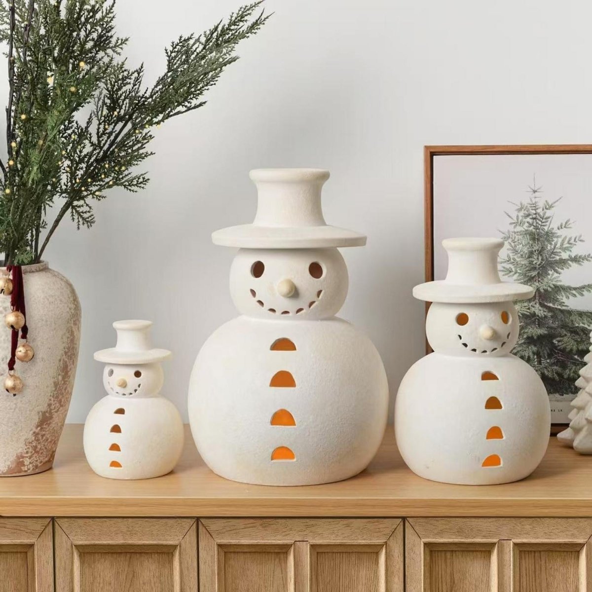 Three minimalist stoneware snowman candle holders displayed on a light wood console table next to a textured vase, showcasing their warm interior glow.