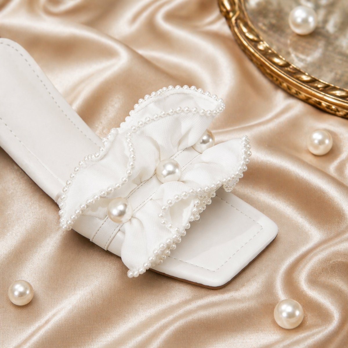 Detailed close-up of a single Kouvr Vionnet white pearl ruffle sandal resting on champagne-colored satin fabric with loose pearls scattered around.