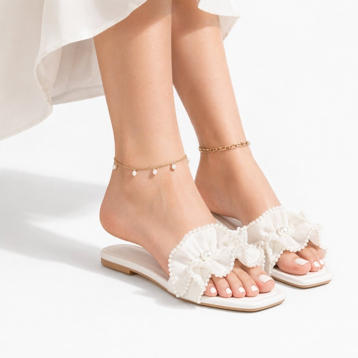 Model wearing Kouvr Vionnet white pearl ruffle sandals with white toenails and two gold anklets, sitting down with a white dress.