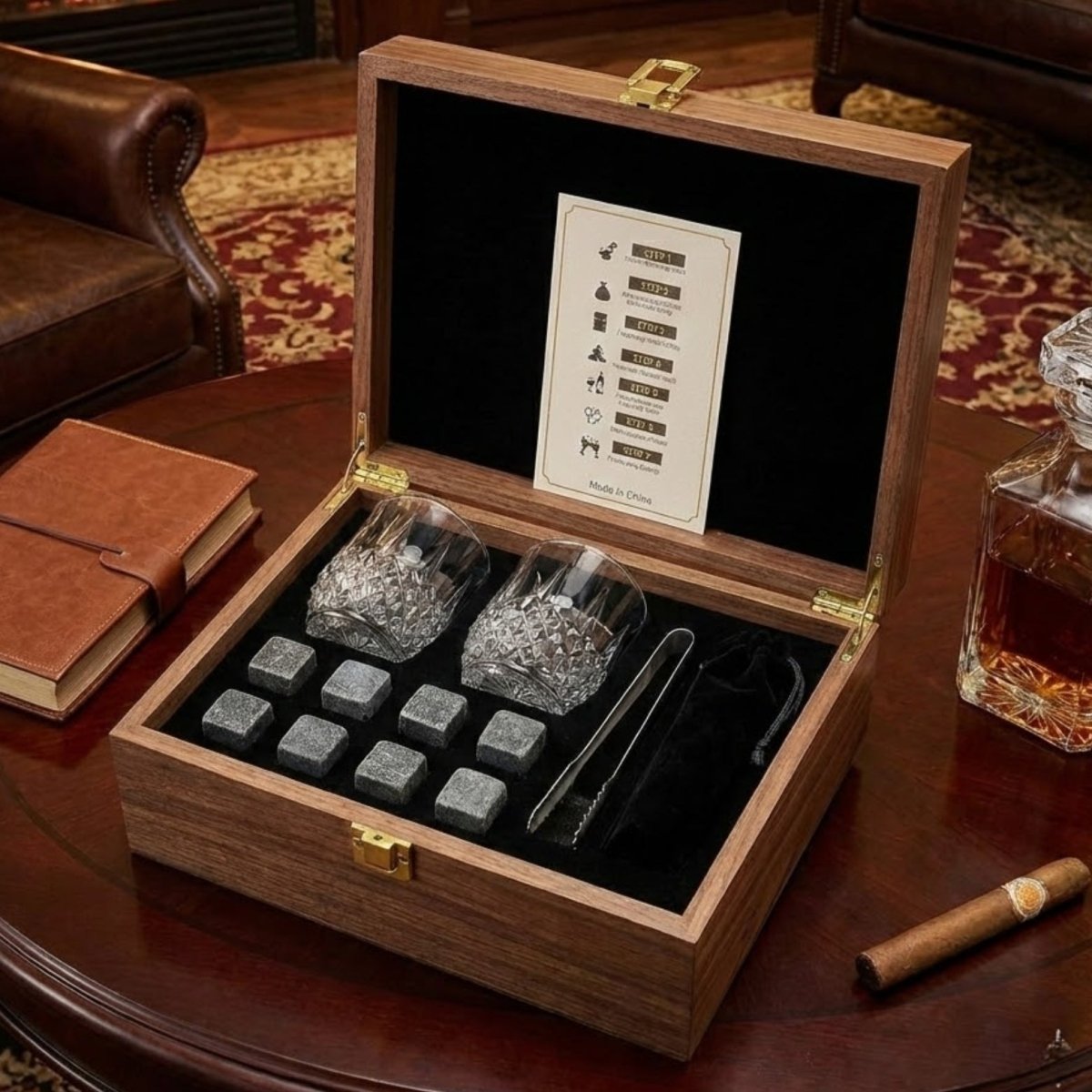 The Kouvr whiskey stones gift set displayed open in its wooden box on a round table, showing two crystal glasses, eight granite stones, and tongs.